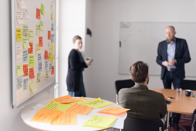 En whiteboard-tavla med massa post-it på, med personer i bakgrunden som håller workshop